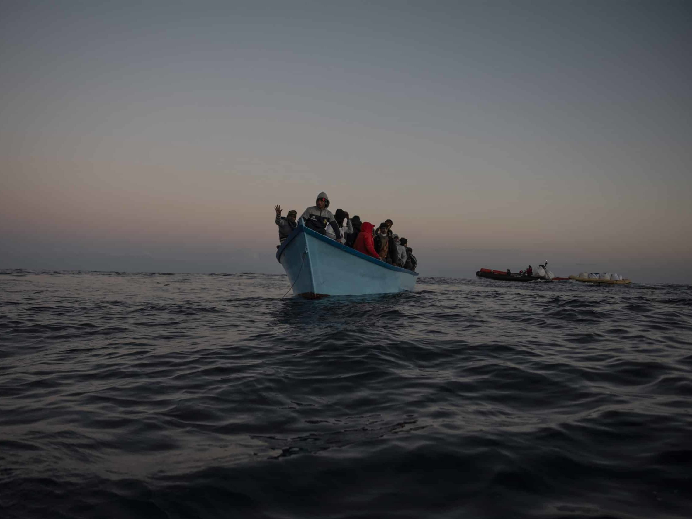 50 Menschen auf blauen Holzboot in Seenot. Im Hintergrund ist die Crew der Sea-Watch 5 mit Rettungsausrüstung zu sehen.