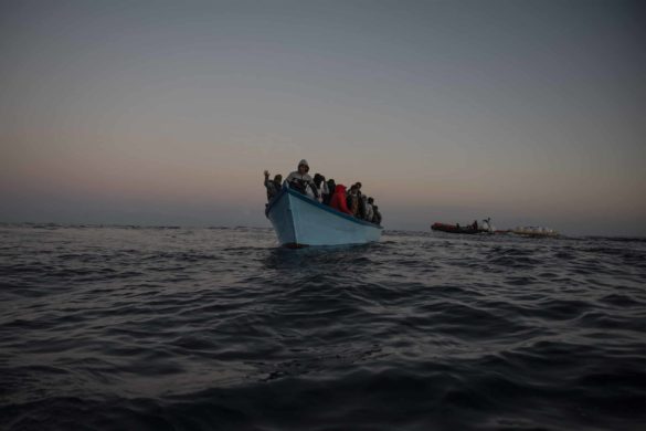 50 Menschen auf blauen Holzboot in Seenot. Im Hintergrund ist die Crew der Sea-Watch 5 mit Rettungsausrüstung zu sehen.