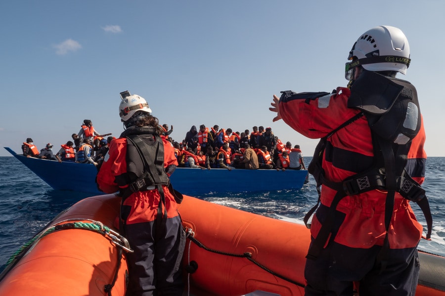Zwei Personen von Sea-Watch befinden sich wenige Meter vor einem Seenotrettungsfall und statten Menschen mit Rettungswesten aus.